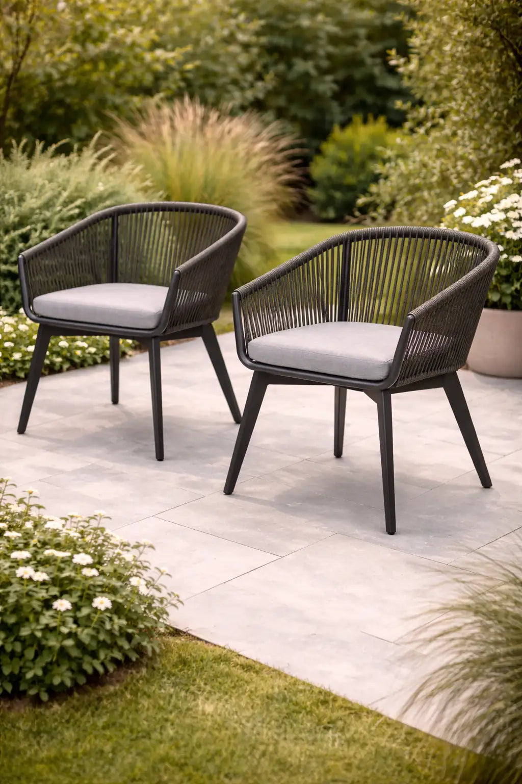 Two black wicker chairs with gray cushions on a stone patio surrounded by greenery.